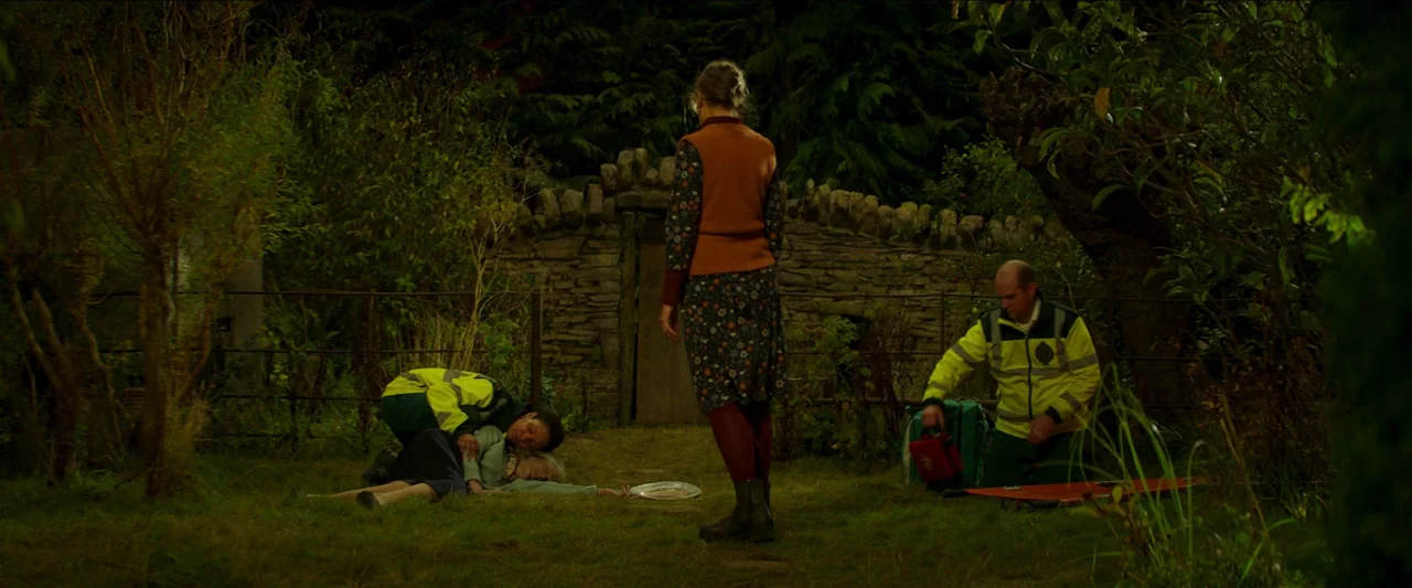 Unwelcome (2022) movie still: wide — A nighttime backyard features a stone wall and dense foliage surrounding the lawn. Two paramedics in…