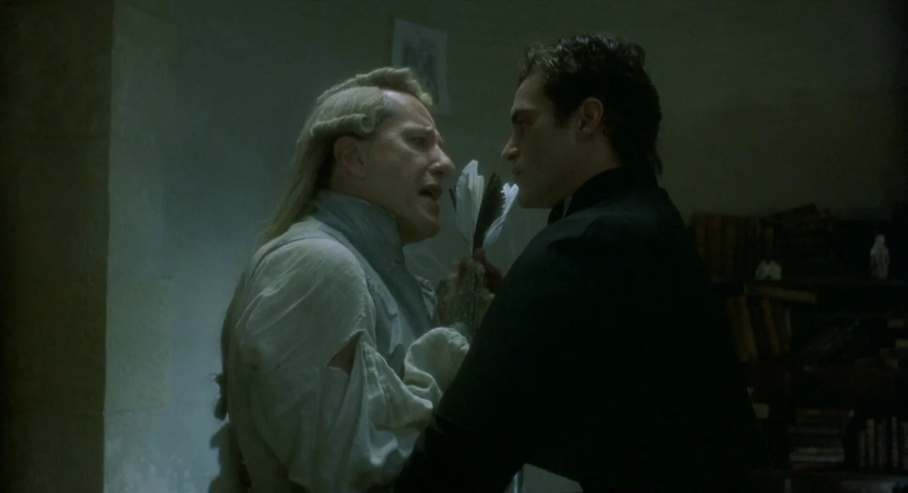 Quills (2000) movie still: medium — A dimly lit room with a bookshelf and plain stone walls. Two men stand close together as one holds quill pens…