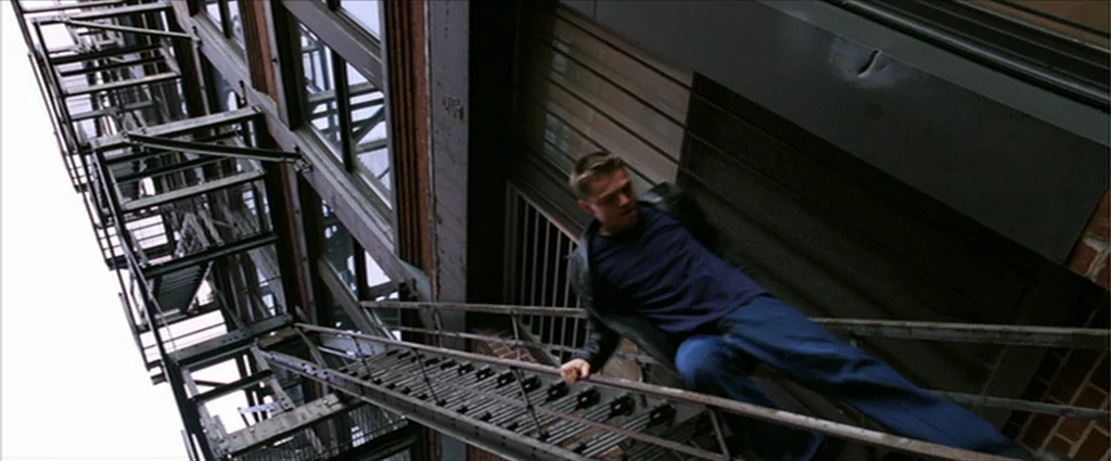 The Departed (2006) movie still: medium — An industrial metal fire escape attached to the side of a red brick building. A young man grips the side…