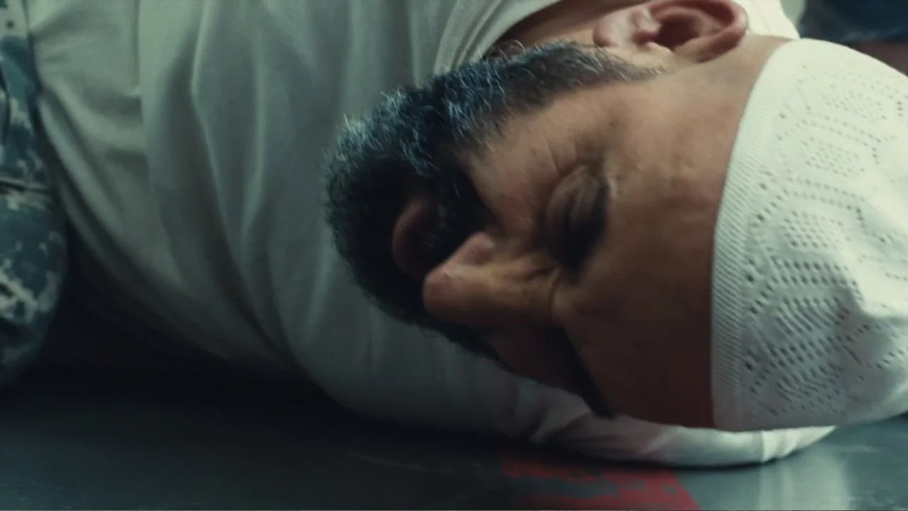 Camp X-Ray (2014) movie still: closeup — An individual lies flat on a smooth surface inside a sterile building. The person wears a white t-shirt and…