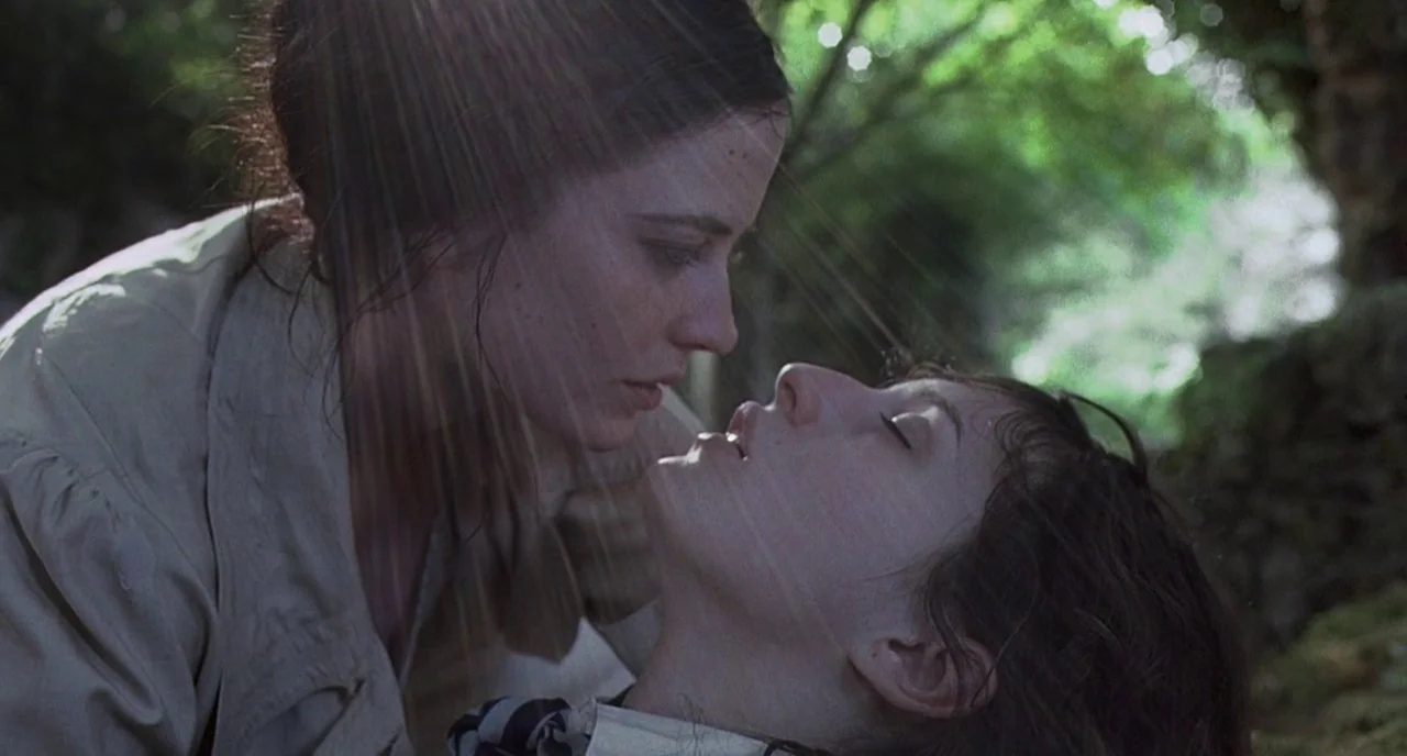 Cracks (2009) movie still: medium — A woman leans over another woman reclining on the ground amidst lush greenery. Strong lens flares streak across…
