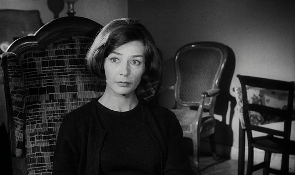Thérèse Desqueyroux (1962) movie still: medium — A dimly lit room contains multiple wooden chairs and dark surfaces. A woman sits in a patterned…