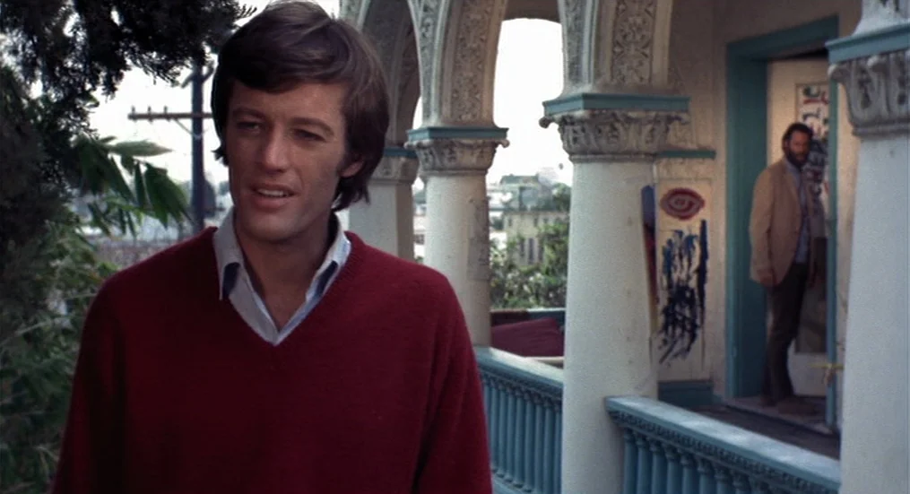 The Trip (1967) movie still: medium — An outdoor balcony features decorative stone columns and a blue painted wooden railing. A young man stands in…
