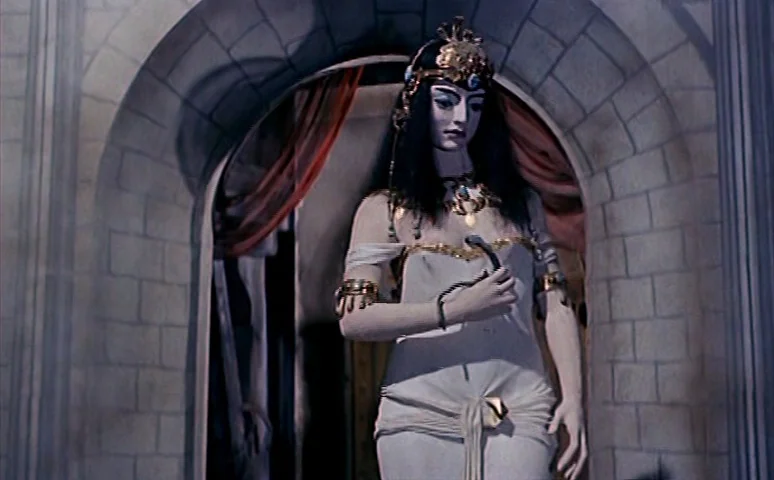 Mill of the Stone Women (1960) movie still: medium — A woman stands in a light stone archway draped with thin red fabric. She holds a thin dark…