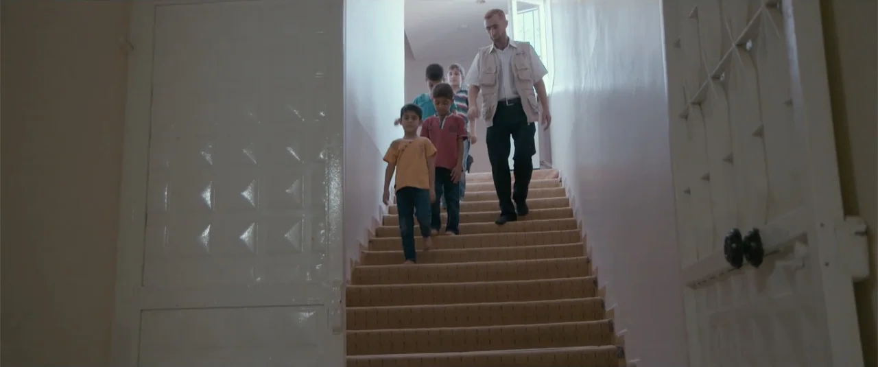 Simple as Water (2021) movie still: fullbody — A residential staircase with beige carpet and plain off-white walls leads to a landing. An adult man…