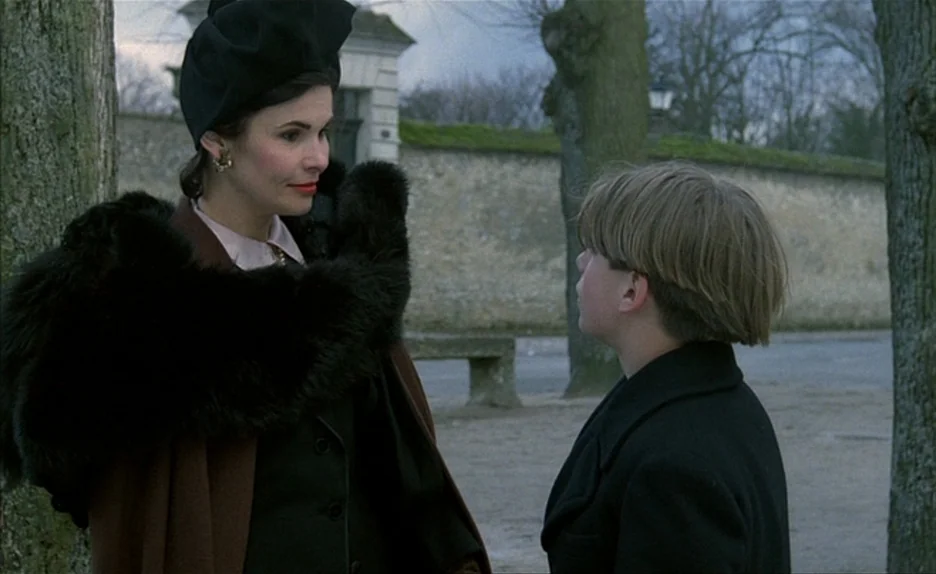 Au Revoir Les Enfants (1987) movie still: medium — A park with mature trees and a stone wall in the background during daytime. A woman in a…