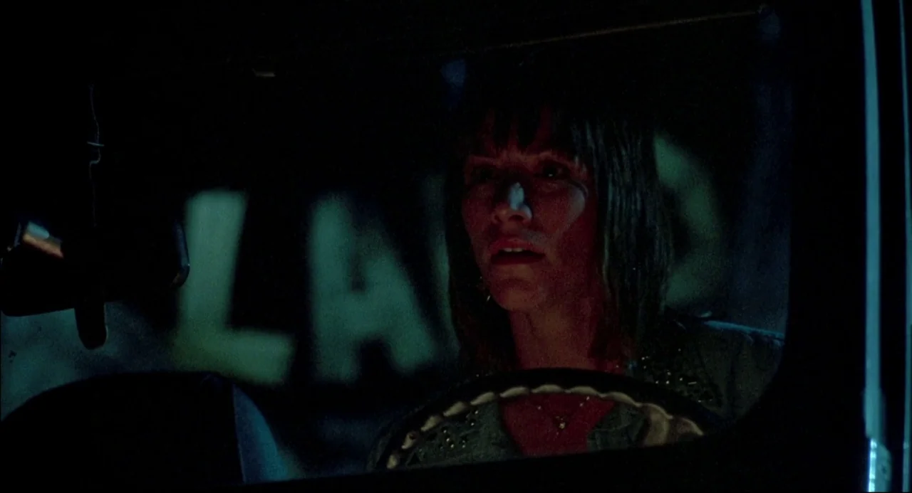 The Texas Chainsaw Massacre 2 (1986) movie still: closeup — A woman sits in the driver seat of a vehicle at night. Green neon light illuminates her…
