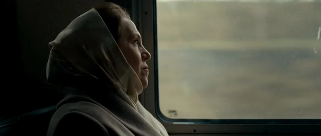 Elena (2011) movie still: closeup — A woman sits inside a train car with a window showing blurred landscape. She gazes out the window as the train…