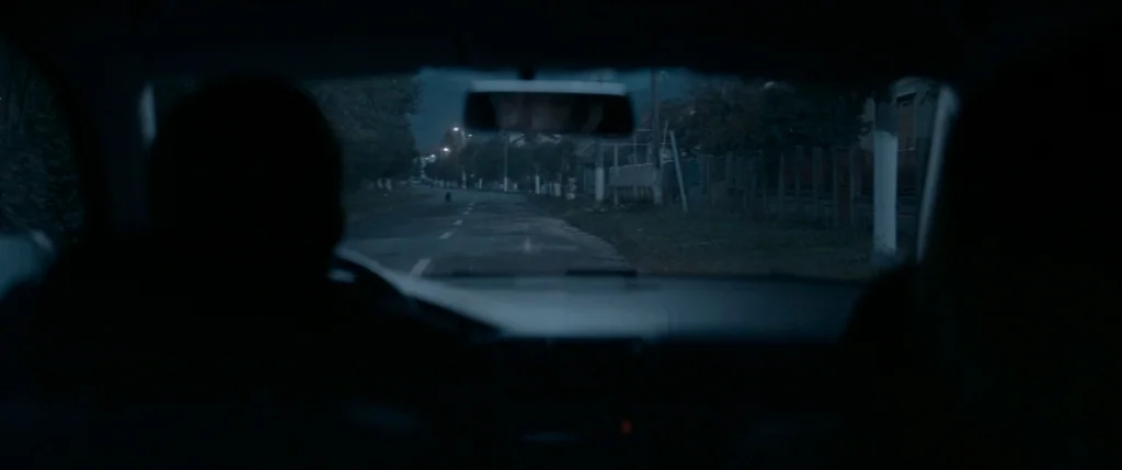Graduation (2016) movie still: medium — A view from inside a car driving along a dark, rural-looking street at night. A driver is silhouetted…