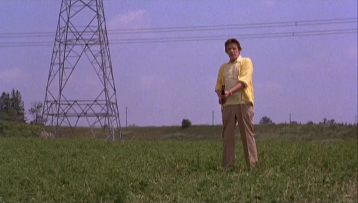 Highway 61 (1991) movie still: fullbody — A grassy field with a large metal electrical tower in the background. A man stands alone holding a small…