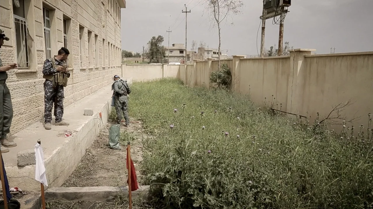 City of Two Springs (2019) movie still: wide — A tan concrete building stands beside a patch of overgrown grass and weeds. Two soldiers in…