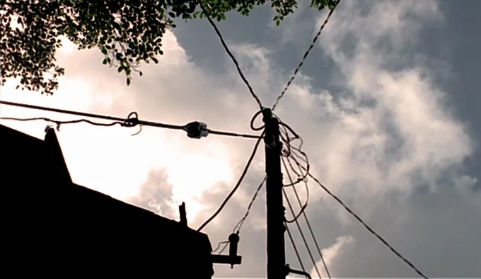 Dust In The Wind (1986) movie still: establishing — A dark utility pole with tangled wires stands against a cloudy, bright daylight sky. Tree leaves…