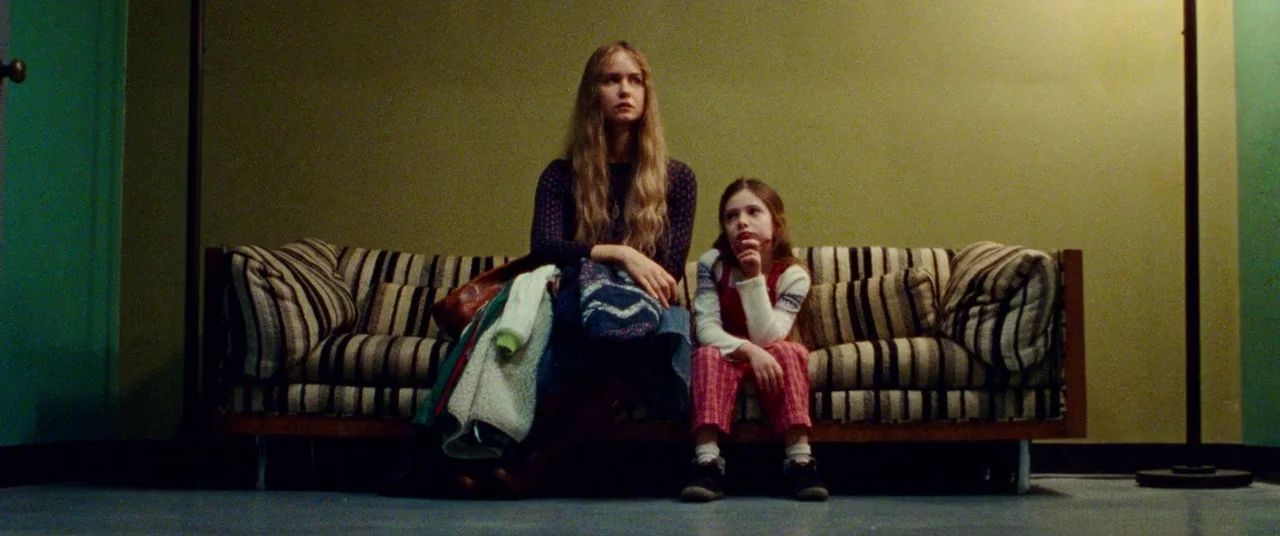 Steve Jobs (2015) movie still: medium — A sparse waiting room with olive green walls and a vintage striped sofa. A young woman and a young girl sit…
