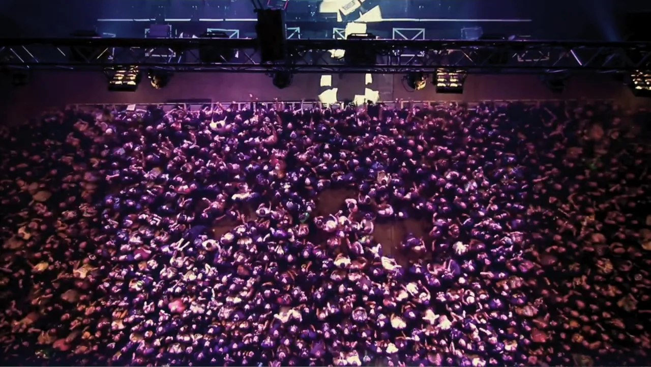 Control (2022) movie still: establishing — A massive concert crowd fills a dark room under bright overhead stage rigging. A multitude of people…