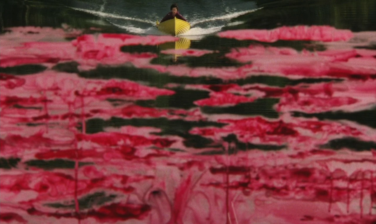 Yumeji (1991) movie still: wide — A dark, reflective lake is covered in swirling patterns of vibrant pink and red. A person sits in a small yellow…