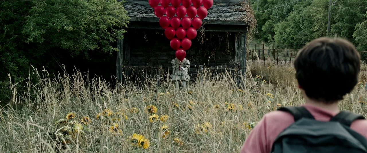 It (2017) movie still: wide — An overgrown field contains dried sunflowers and tall grasses in front of a shed. A person in a white clown costume…