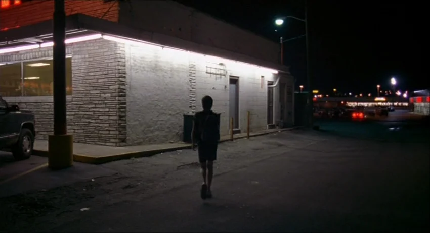 Gummo (1997) movie still: A solitary figure walks away from the camera down a dimly lit street or parking lot at night, their back to the viewer.…