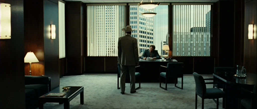 No Country For Old Men movie still: wide — A modern executive office features wood-paneled walls, a large desk, and floor-to-ceiling windows. A man…