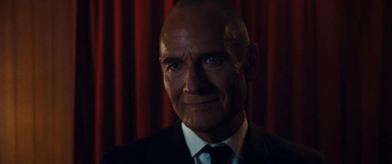 MaXXXine (2024) movie still: closeup — A man stands in front of heavy, dark red fabric curtains. He gazes forward while wearing a formal dark suit…