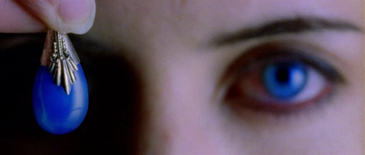 The Strange Colour of Your Body’s Tears (2013) movie still: closeup — A close-up reveals a bright blue pendant held by fingers next to a human eye.…