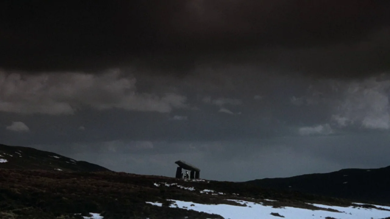 Excalibur (1981) movie still: wide — A remote, hilly terrain covered in patches of snow sits under heavy storm clouds. A solitary megalithic stone…