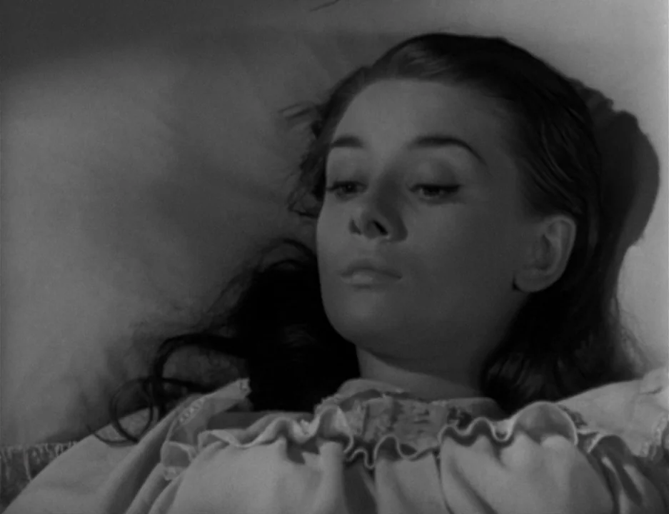 Roman Holiday (1953) movie still: closeup — A person lies in a bed against a white pillow in dim lighting. The subject faces slightly away from the…
