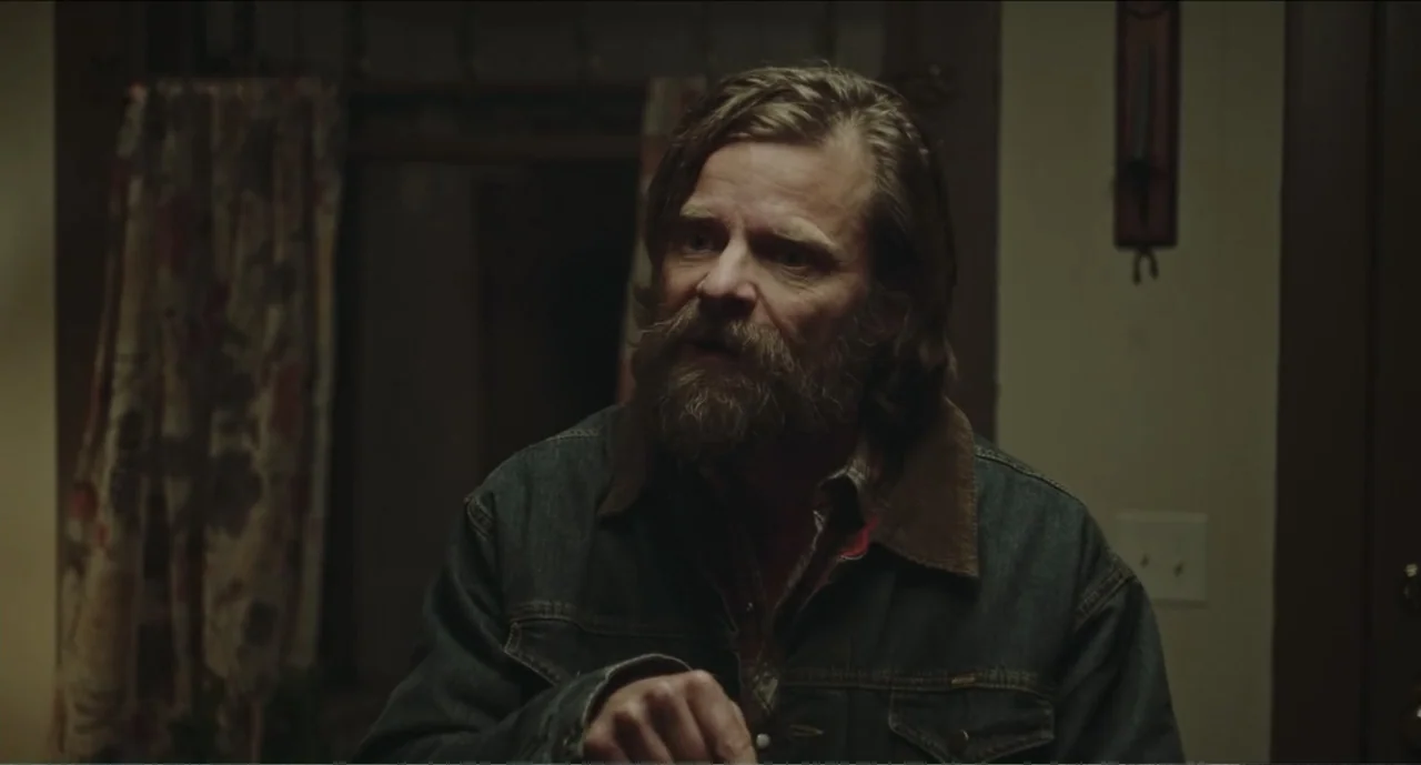 Cowboys (2014) movie still: medium — A dimly lit interior space with a curtain in the blurred background. A man with a full beard and denim jacket…