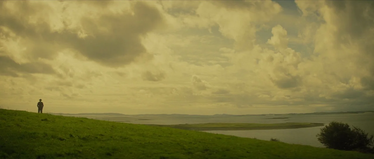 The Lost City of Z (2016) movie still: wide — A vast coastal landscape under a thick, overcast sky with rolling green grass hills. A lone man in a…