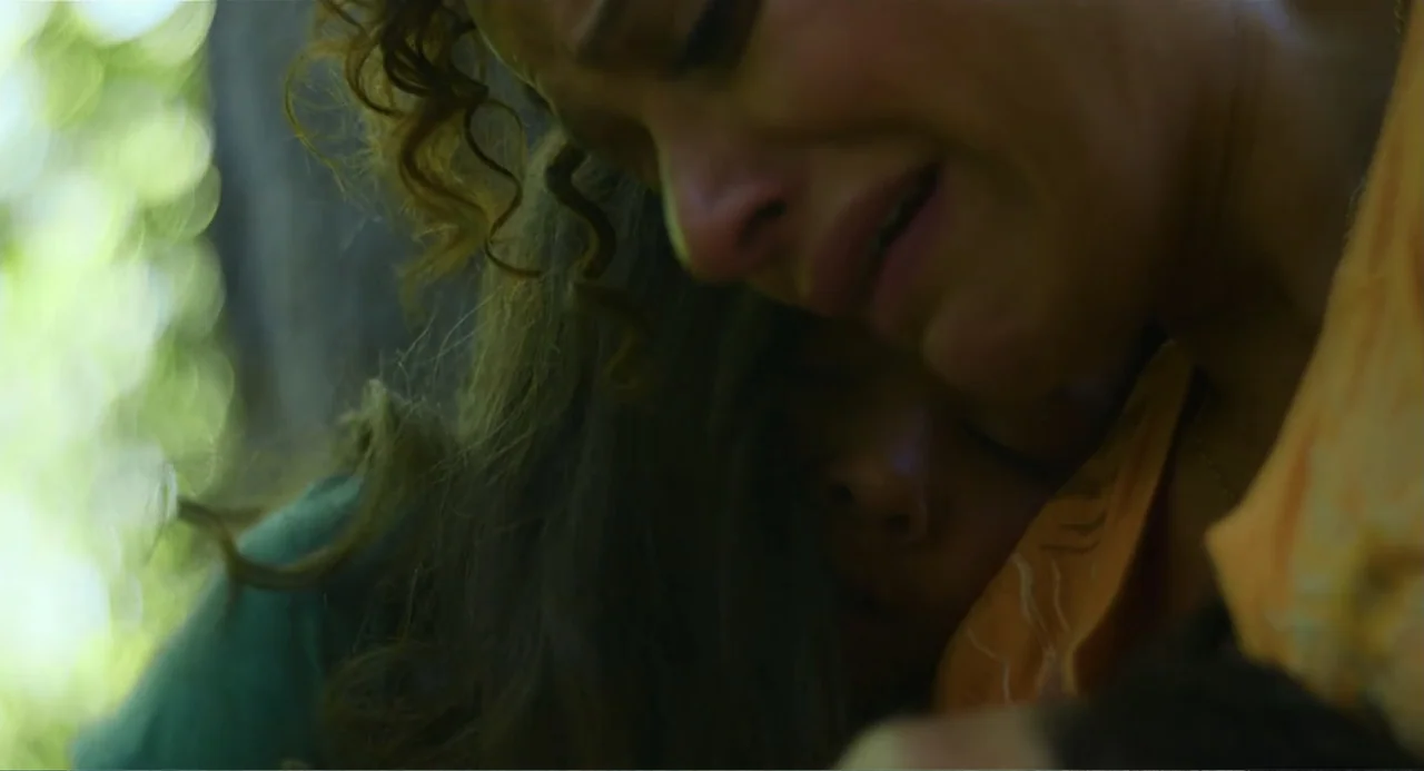 Turtles All the Way Down (2024) movie still: closeup — Two people are embraced in a wooded area with blurred foliage in the background. A woman with…