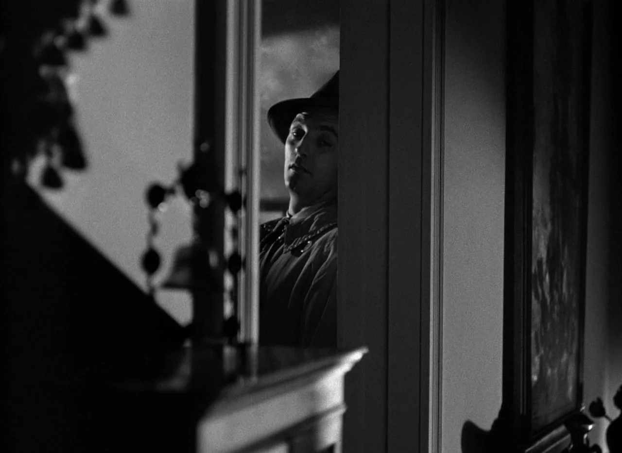 Out of the Past (1947) movie still: medium — A dark, dimly lit domestic interior is framed by the edges of a doorway. A man wearing a fedora stares…