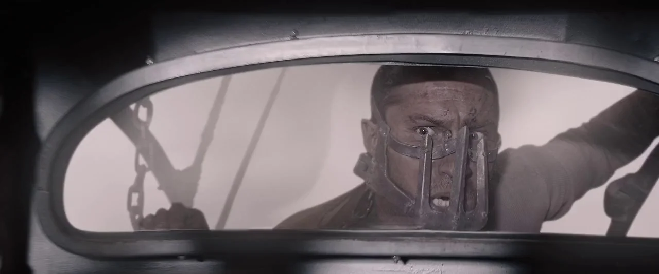 Mad Max: Fury Road (2015) movie still: closeup — A man looks through a dirty vehicle rear window in a desert wasteland. He wears a heavy metallic…