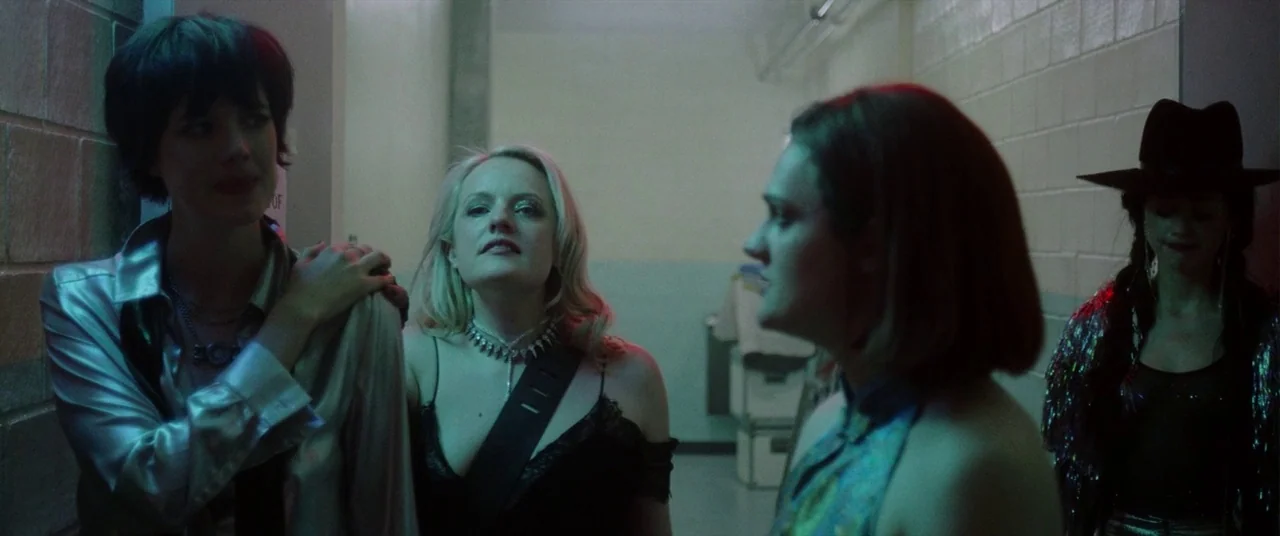 Her Smell (2018) movie still: medium — A narrow industrial passage features beige concrete block walls and harsh overhead fluorescent lights. Four…