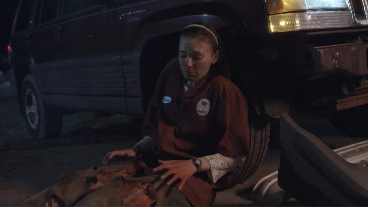 Burn (2019) movie still: medium — A dark night setting beside a parked vehicle in a dimly lit area. A young woman in a work uniform sits on the…