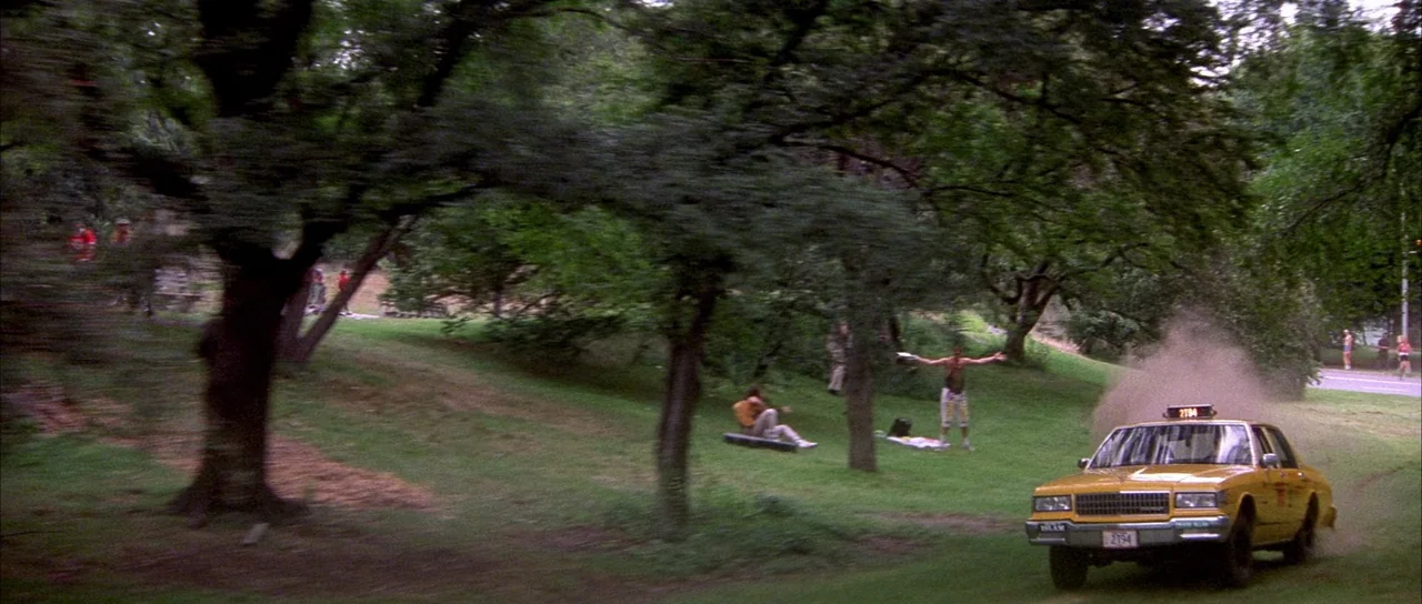 Die Hard with a Vengeance (1995) movie still: wide — A green public park features numerous mature deciduous trees and sloped grassy terrain. A…