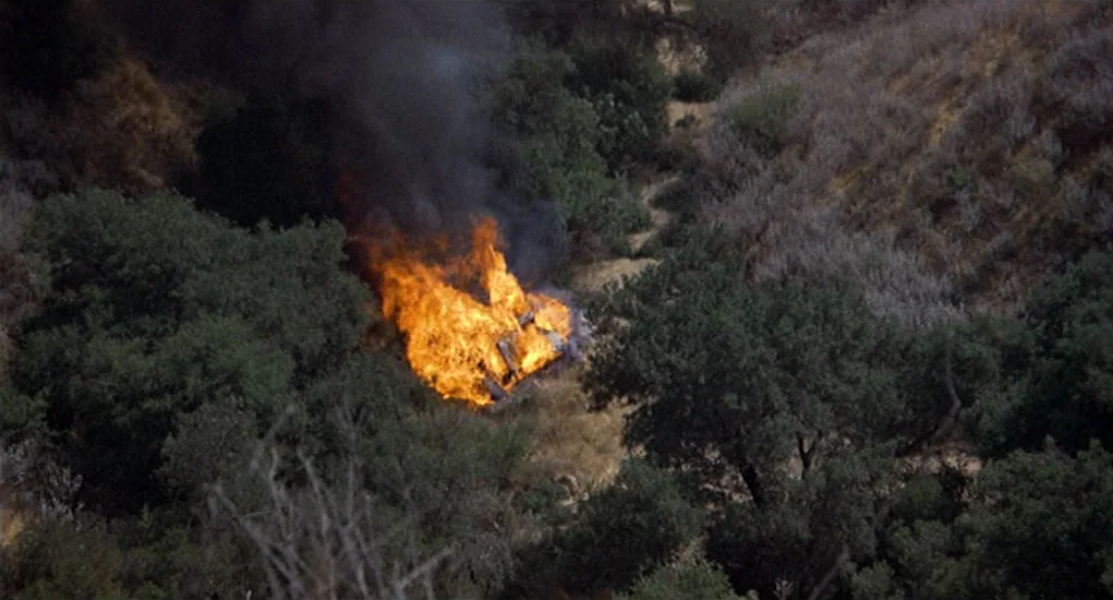 Family Plot (1976) movie still: wide — A burning car sits in a dry, scrubby canyon filled with brush and trees. Intense orange flames and thick…