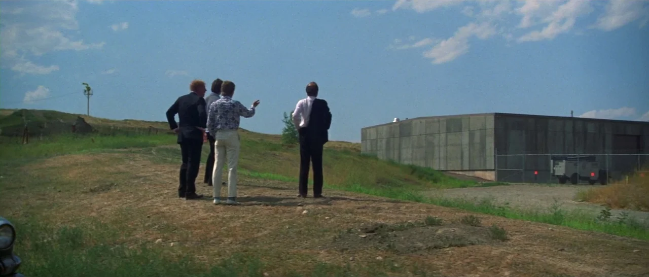 Thunderbolt & Lightfoot (1974) movie still: wide — Three men stand on a barren dirt hill beside a flat concrete warehouse building. The sky is clear…