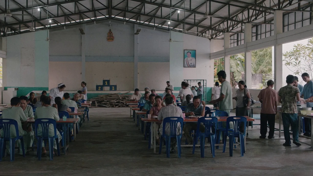 Cemetery of Splendour (2015) movie still: wide — A large, open-air communal dining space with a metal truss roof and concrete floors. Many people…