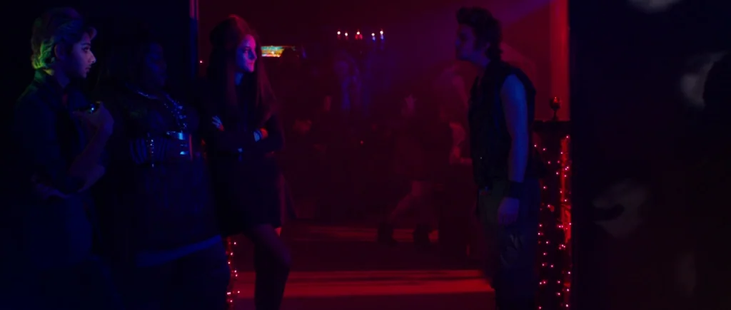 White Bird in a Blizzard (2014) movie still: wide — A dark, dimly lit nightclub interior filled with deep red and blue neon light. A group of three…