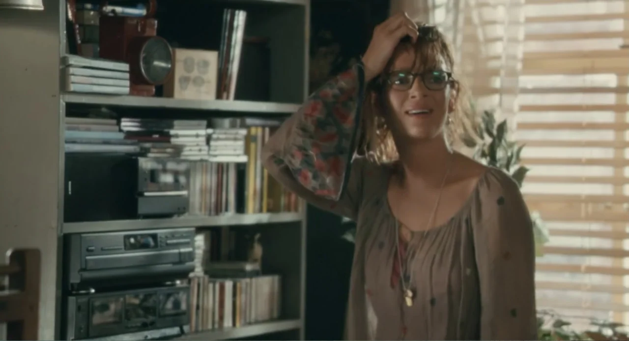 Motherhood (2020) movie still: medium — A cluttered interior living space features a shelf unit packed with books and audio equipment. A young woman…