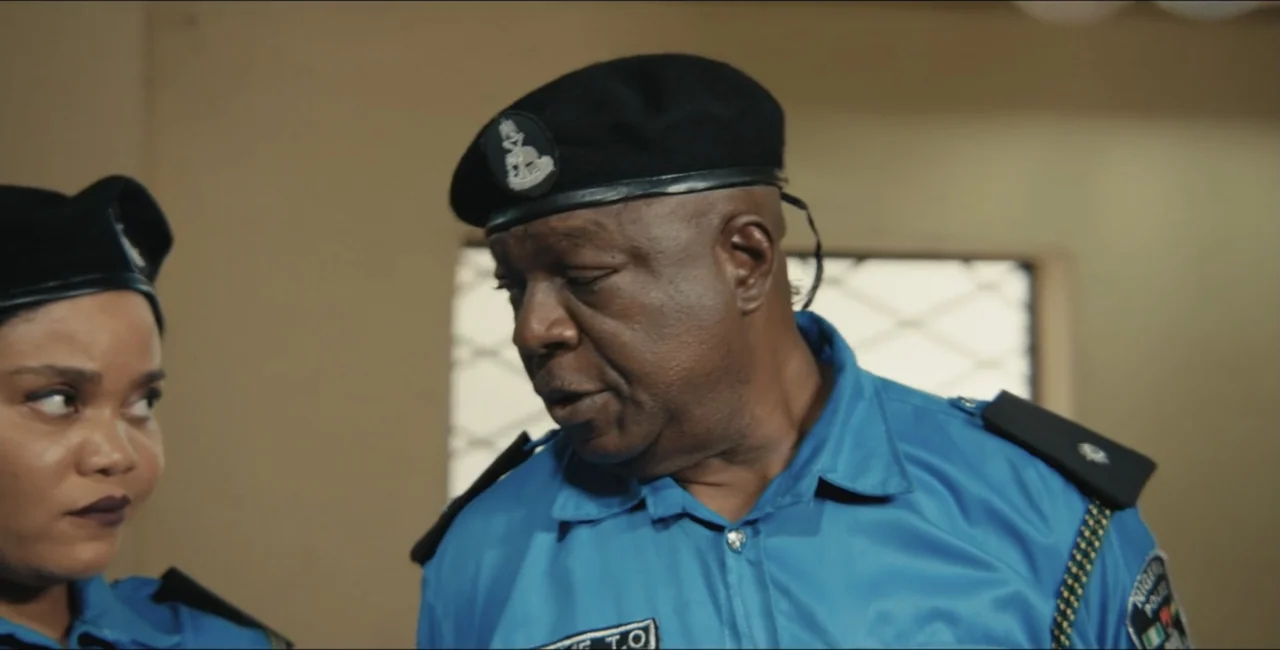 Project Assault (2025) movie still: medium — A room with plain beige walls and a window with a metal grid. Two uniformed police officers stand close…