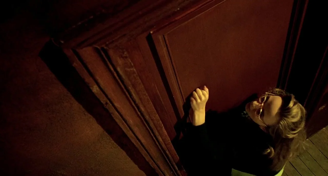 Delicatessen (1991) movie still: medium — A woman stands in a dark corner outside a large, dark-wood door at night. She raises her hand to knock on…
