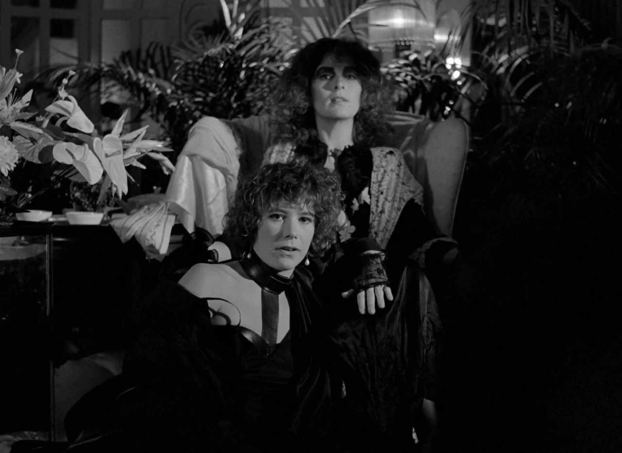Singapore Sling (1990) movie still: medium — A dimly lit room filled with large potted palm leaves and artificial flowers. Two women sit together…