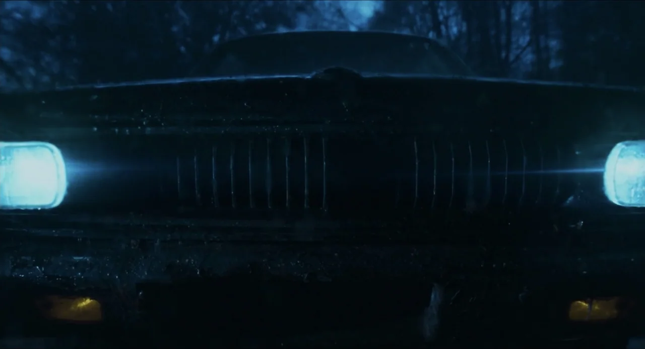 Echoes Of A Crime (2025) movie still: closeup — The front grill and glowing headlights of a car dominate the dark frame. It is night and the vehicle…