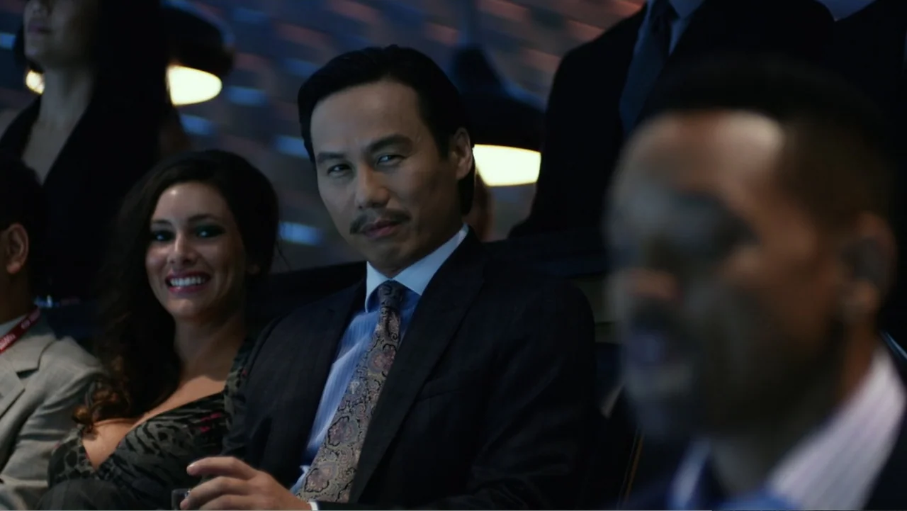 Focus (2015) movie still: medium — A dimly lit luxury viewing suite with multiple rows of plush seating. An East Asian man in a suit sits among…