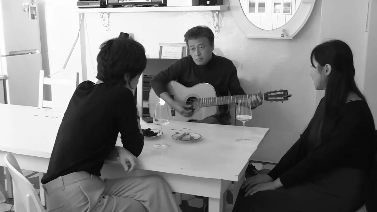 Walk Up (2022) movie still: medium — Three people sit around a white dining table in a simply decorated, monochromatic room. The man in the center…