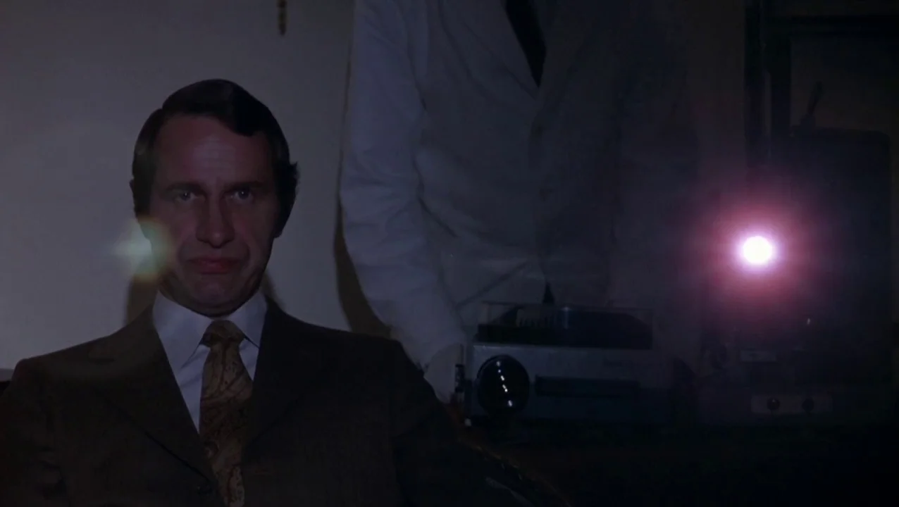 O Lucky Man! (1973) movie still: medium — A dark indoor setting features a seated man facing forward in a dimly lit room. Beside him a person in a…