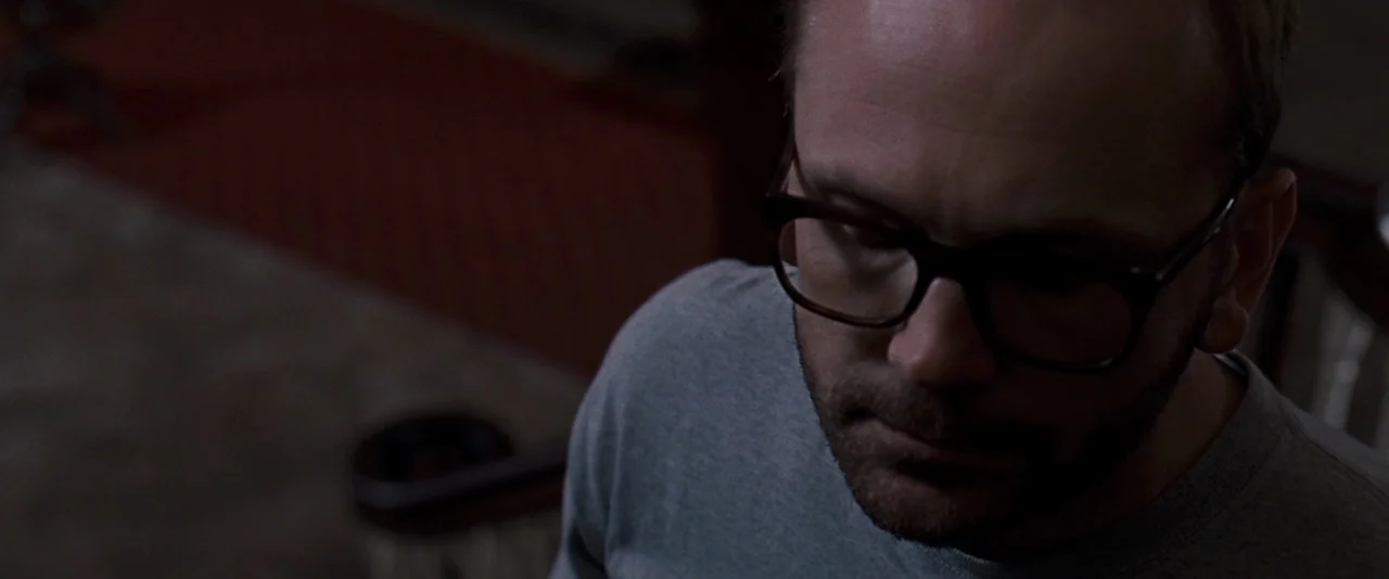 The Innkeepers (2011) movie still: closeup — A man stands in a dark, interior space with a visible banister in the background. The man looks down…