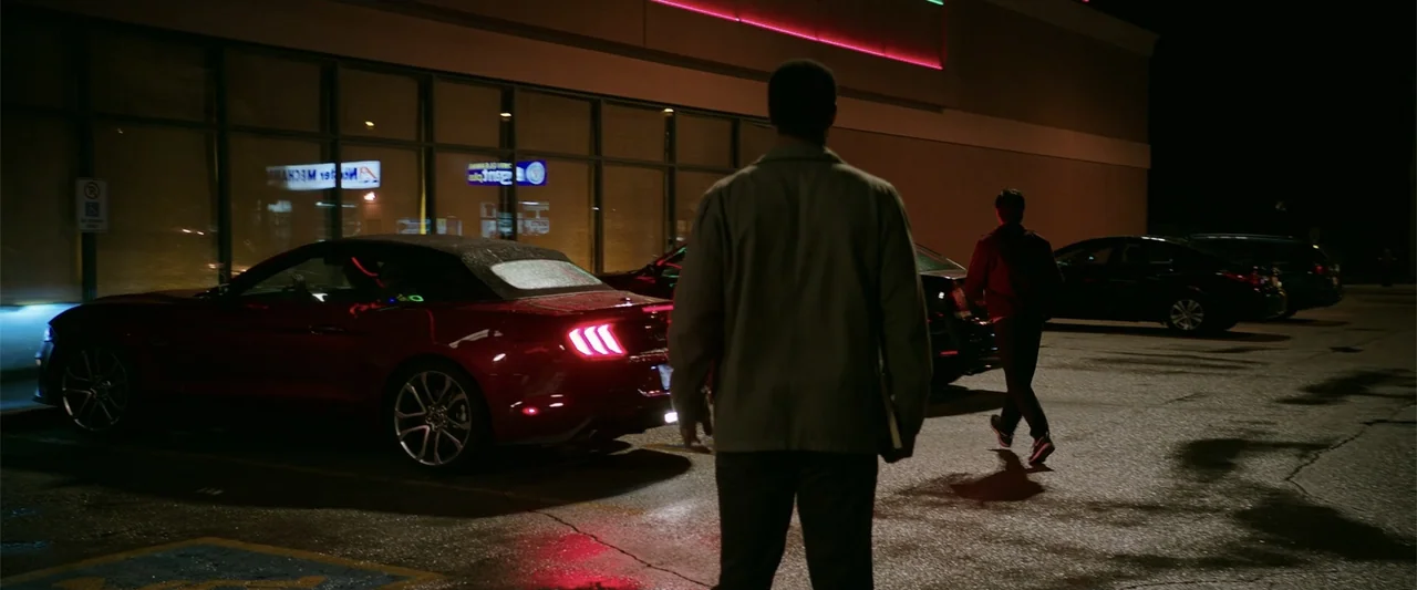 K.G.F: Chapter 2 (2022) movie still: fullbody — A nighttime parking lot exterior features two men standing near a parked red car. One man is…