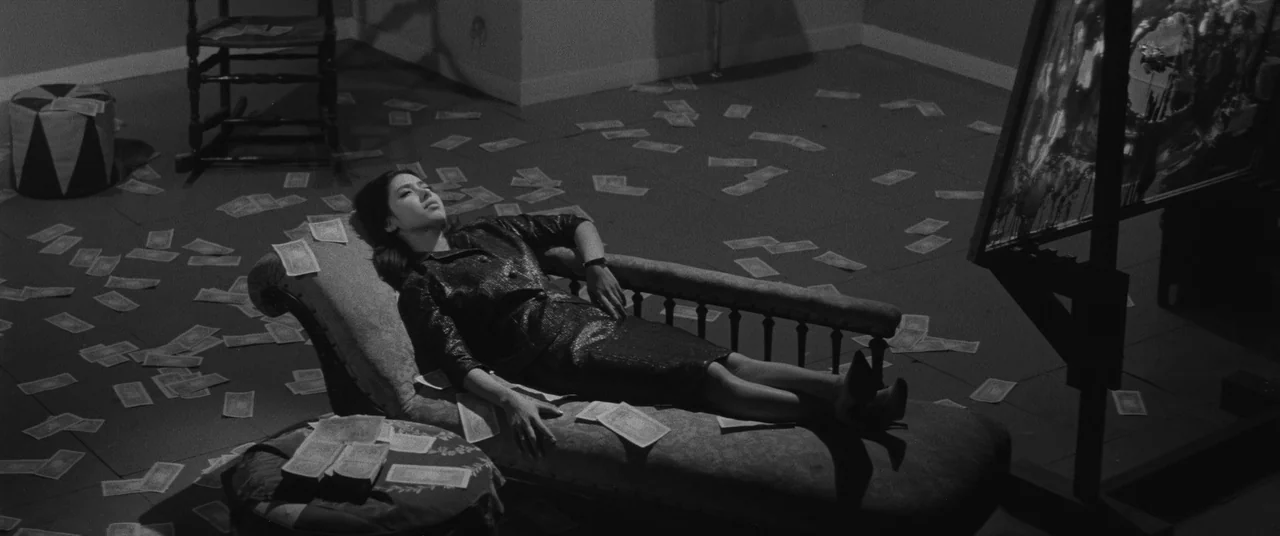 Carmen From Kawachi (1966) movie still: wide — A spacious room features a dark floor covered in scattered paper banknotes. A young woman rests on a…