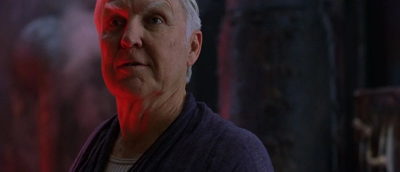 The Matrix Reloaded (2003) movie still: closeup — A dark industrial space features metallic textures bathed in a sharp red glow. An elderly man with…